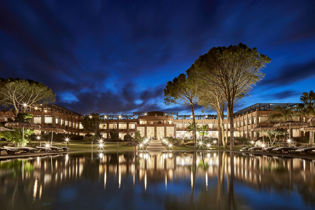Hotel gardens and illuminated buildings reflected in a swimming pool beneath a deep blue evening sky in Sardinia